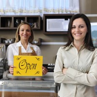 שתי נשים מחייכות עומדות בחנות כשאחת מהן מחזיקה שלט צהוב עם הכיתוב "OPEN"