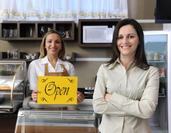 שתי נשים מחייכות עומדות בחנות כשאחת מהן מחזיקה שלט צהוב עם הכיתוב "OPEN"